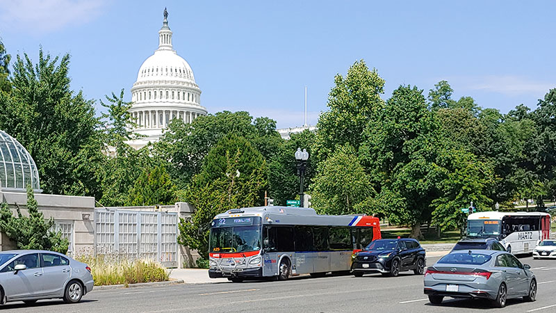 Photo of a DC bus traveling with other car traffic on Constitution Ave NE in Washington, DC with the US Capitol in the background.